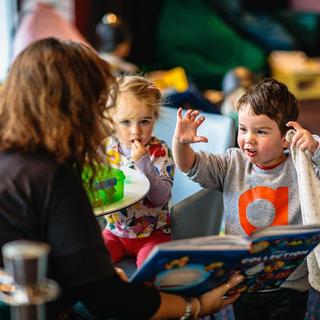 A woman reads a story book to two small children; a boy raises his hands as if roaring. Toys are in the background.