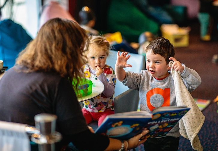 A woman reads a story book to two small children; a boy raises his hands as if roaring. Toys are in the background.