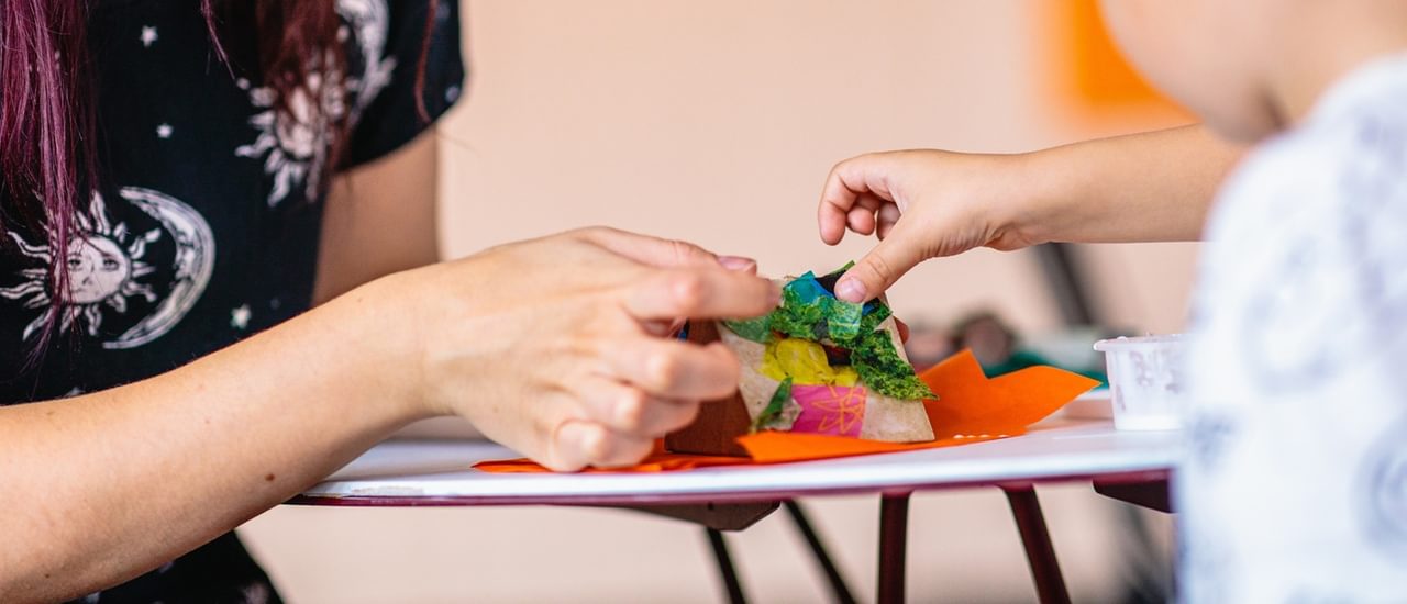 A close up of a woman and boy are sticking a gluing colourful pieces of tissue paper to a model.