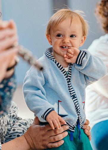 A toddler in a blue cardigan smiles with his hand up to his mouth. He is looking towards a musician playing the flute. Only the end of the flute and the musician's hands can be seen.