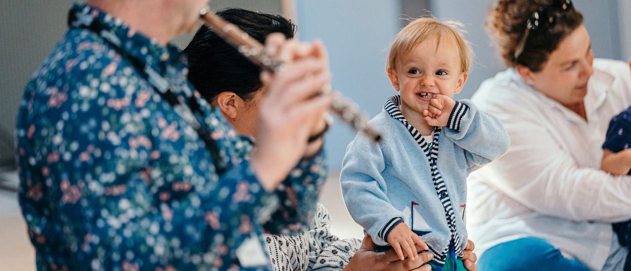 A toddler in a blue cardigan smiles with his hand up to his mouth. He is looking towards a musician playing the flute. The muscian's face is obscure, he is wearing a spotty blue shirt.
