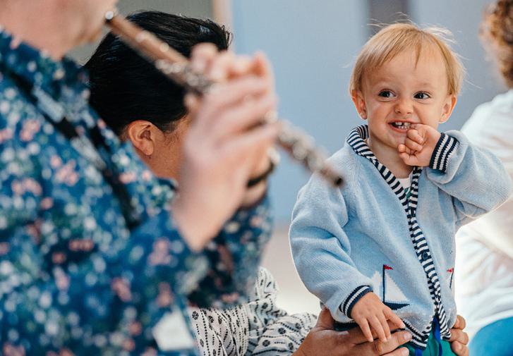A toddler in a blue cardigan smiles with his hand up to his mouth. He is looking towards a musician playing the flute. The muscian's face is obscure, he is wearing a spotty blue shirt.