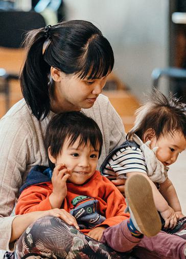 A woman with tied back dark hair holds a toddler and a baby, they are all smiling. The woman wears a beige jumper, the toddler on the left wears an orange hoodie, and the baby wears blue dungarees and a stripy shirt.