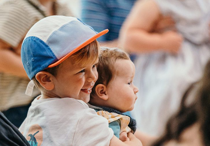 A young boy in a blue baseball cap smileswhile holding a baby. Next to the boy a woman in a black shirt, with a gold earing and white hair tie smiles.