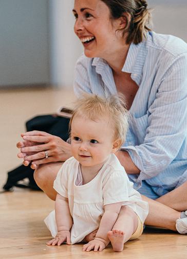 A woman in a blue shirt sits on a wooden floor smiling, with her hands together. In front of her sits a blond baby in a white top, also smiling.