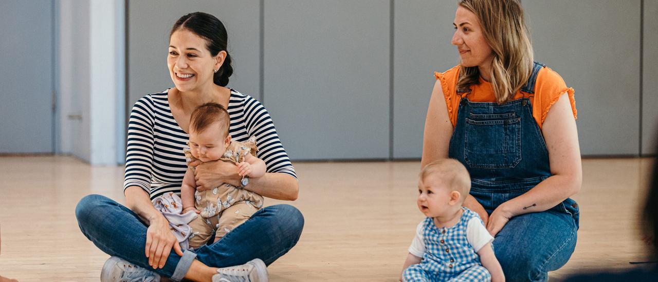 On the left a young woman with black hair in a striped T-shirt and jeans sits cross legged on a wooden floor, holding a baby and smiling. On the right a blond woman in dungarees and an orange shirt kneels. A baby wearing a blue sits in front of her.