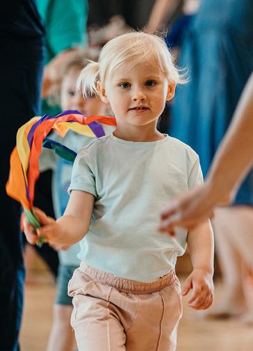 A child with blond hair in a blue t-shirt smiles at the camera. They are holding a mutlicoloured ribbon. Other children and parents can be seen, blurry, in the background.