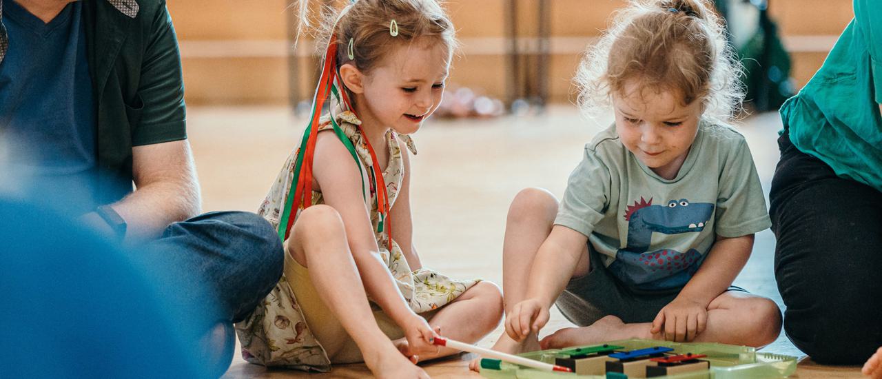 Two young girls sit cross legged on the floor, playing a glockenspiel and smiling. The girl on the left has red and green ribbons in her hair, the girl on the right has a dinosaur on her t-shirt.