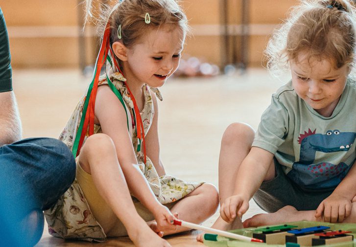 Two young girls sit cross legged on the floor, playing a glockenspiel and smiling. The girl on the left has red and green ribbons in her hair, the girl on the right has a dinosaur on her t-shirt.