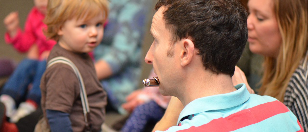 A boy in a brown T-shirt and dungarees watches a man in a stripey pink and blue shirt play the flute