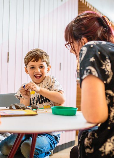A child and their parent are sat playing with crafts at a table and smiling at the camera.