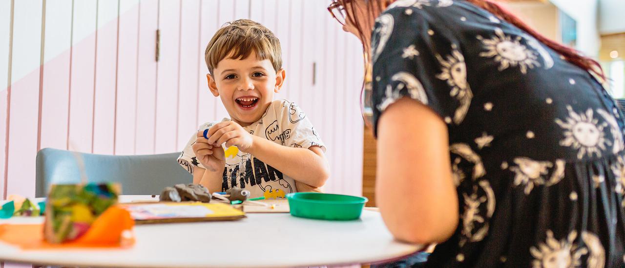 A child and their parent are sat playing with crafts at a table and smiling at the camera.