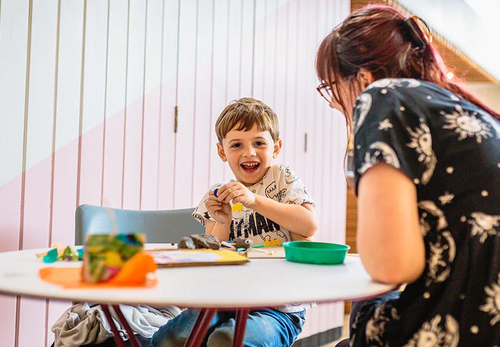 A child and their parent are sat playing with crafts at a table and smiling at the camera.