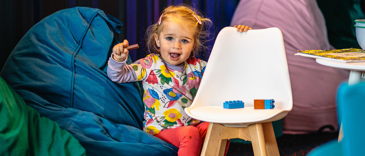 A toddler sits on a beanbag in the foyer of the Theatre. They are holding a crayon.