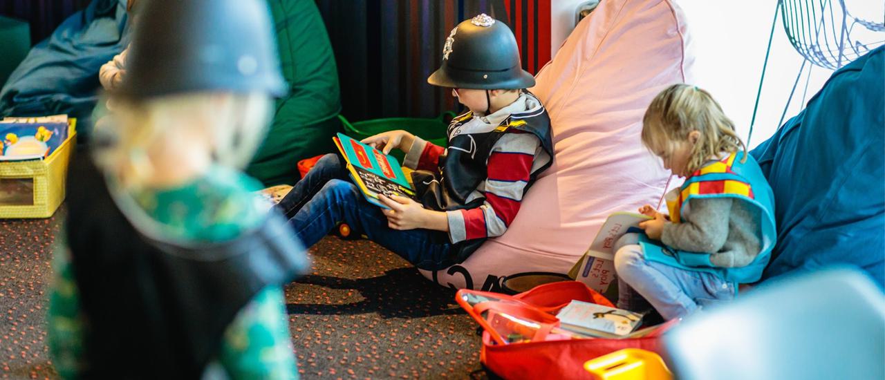 Children in fancy dress outfits and hats sit on beanbags, reading.