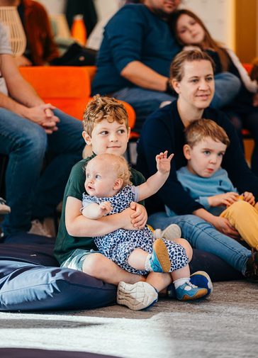 A little boy sits, with a baby on his lap, intently listening to a story; sat on beanbags and chairs.