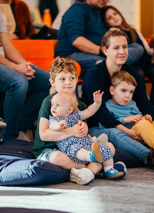 A little boy sits, with a baby on his lap, intently listening to a story; sat on beanbags and chairs.