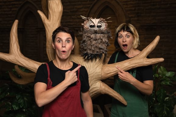 Two ladies wearing brightly coloured dungarees and looking shocked pinting at a tree where a puppet owl sits.