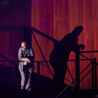 An reddish brown backdrop with a black metal staircase platform with stairs in front. To the left a man in a grey shirt and trousers leans over the railings, looking to the left. His dark shadow looms large on the backdrop behind.