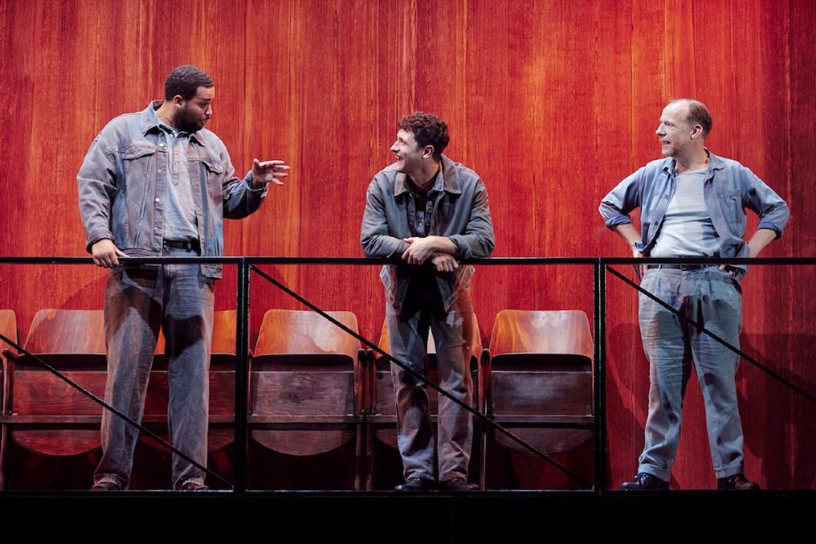 Three men, in grey trousers, grey shirts and black boots, lean over a black metal railing. The man on the left is speaking to the other two and gesticulating with his left hand; the other two are looking at him and smiling. Behind them is a row of wooden fold up seats, and a reddish brown wooden backdrop.