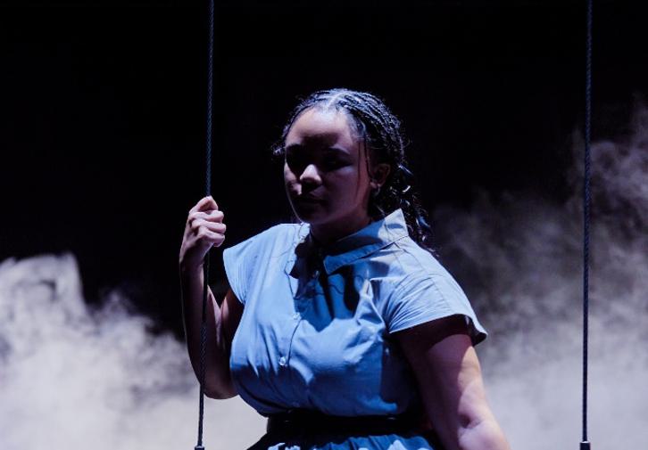 A woman in a light grey dress sits on a swing. Her face is in the shadows and behind her the stage is dark, with swirling grey smoke.