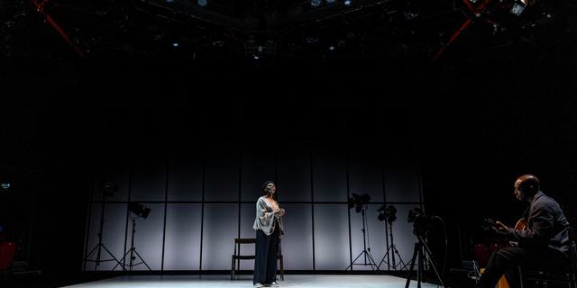 Rakie (playing Adrienne) and Jack (playing Adam) are standing across from each other on a wide open stage. The floor is a bright white, behind them is a panelled grey wall and positioned around the space are stage lights. Jack is listening intently as Rakie speaks out to the audience.