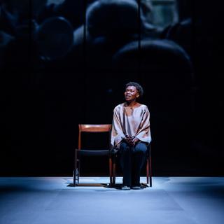Rakie (playing Adrienne) is seated on one of two chairs in the middle of the empty stage. Projected onto the wall behind her is a blurry photo of a crowd, as if appearing from her memory.