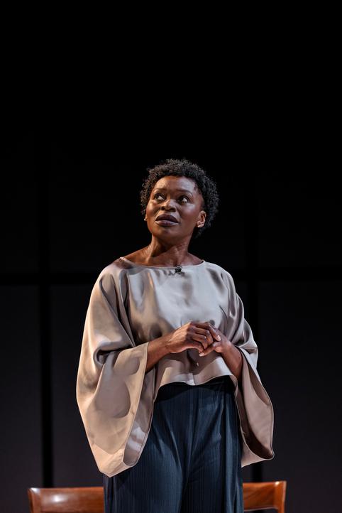 Rakie (playing Adrienne), wearing a silver-coloured, flowing blouse and wide-legged dark trousers is looking out towards the audience with a pensive look on her face.