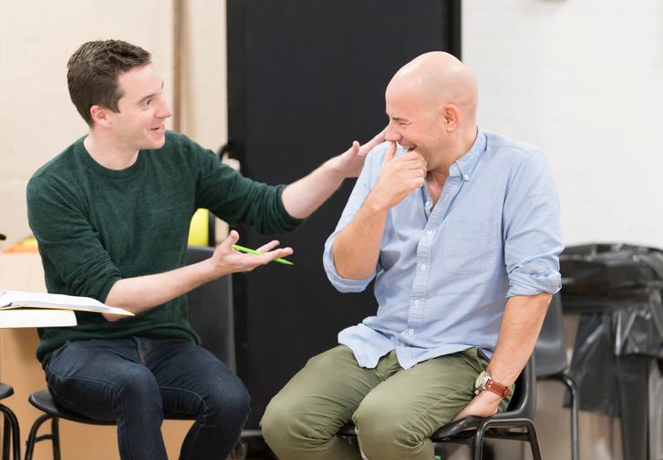 James Graham and Daniel Evans sit next to each other. James, with short dark hair, wears a dark green turtleneck jumper and is raising his arms, gesturing towards Daniel who has his hand to his mouth, laughing. He's wearing a pale blue shirt and green trousers.
