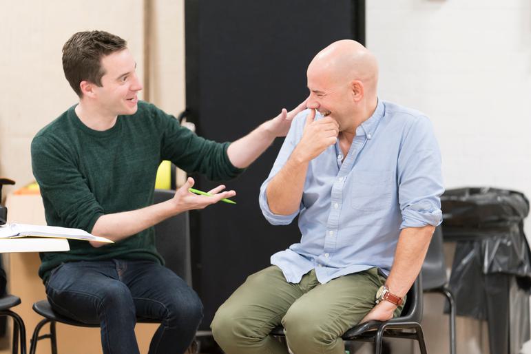 James Graham and Daniel Evans sit next to each other. James, with short dark hair, wears a dark green turtleneck jumper and is raising his arms, gesturing towards Daniel who has his hand to his mouth, laughing. He's wearing a pale blue shirt and green trousers.