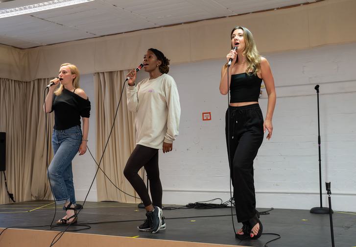 Carly Bawden, Angela Marie Hurst and Zizi Strallen singing on a raised platform in front of a white wall with cream curtains. The are all standing with a microphone in thier right hand and their left knee slightly bent.
