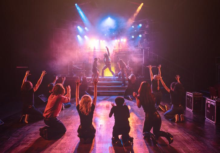 A performer on stage backlit by bright coloured lights with company members gathered round in a circle, kneeling with their arms in the air towards the stage.