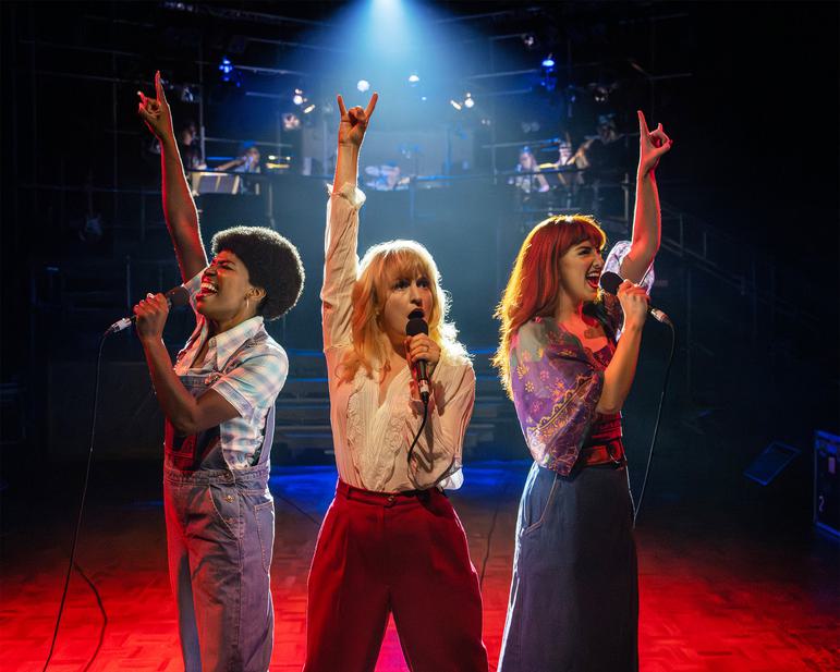 Dee (Angela Marie Hurst), Anna (Carly Bawden) and Q (Zizi Strallen) stand wearing typically 70s clothing, they all hold a microphone in one hand and have there other arm raised above their heads making a rock sign. they are lit by one main spotlight.