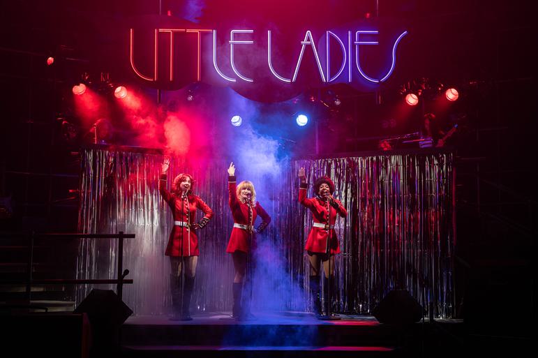 Q (Zizi Strallen), Anna (Carly Bawden) and Dee (Angela Marie Hurst) are dressed in red dresses that resemble Royal Guard outfits. They are standing on a stage that is lit with red, white and blue lights, they all have one arm in the air. Above them, lit up letters in red, white and blue read 'Little Ladies'.