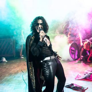 Stevie (sebastien Torkia) is dressed in black leather trousers, a black cape and black top. He has long black curly hair and is leaning back singing into a microphone. Behind him is a lot of coloured smoke and haze and in the background some dustbins with people inside them.
