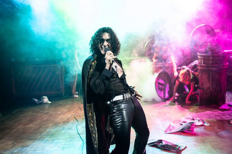 Stevie (sebastien Torkia) is dressed in black leather trousers, a black cape and black top. He has long black curly hair and is leaning back singing into a microphone. Behind him is a lot of coloured smoke and haze and in the background some dustbins with people inside them.