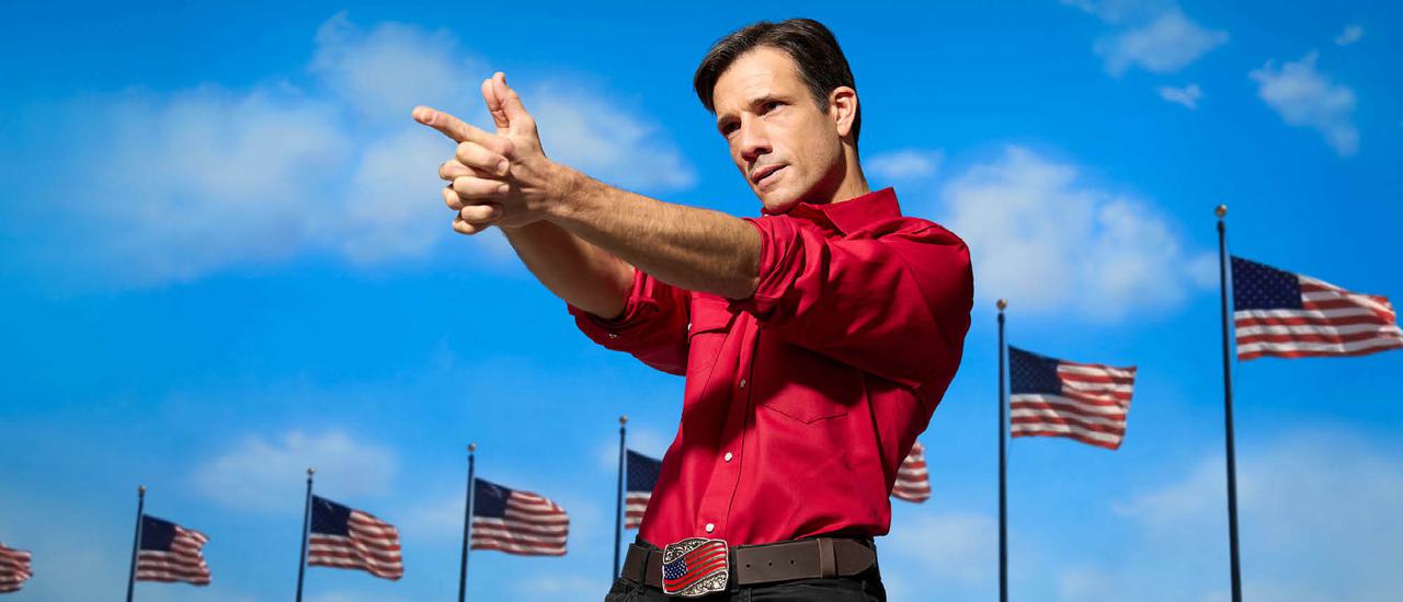 A man (Danny Mac), wearing a red shirt, black jeans and a belt with a US Stars & Stripes flag on the buckle, stands with his arms and hands pointing ahead of him as though firing a gun. Behind him is a brilliant blue sky and a row of American flags flying at full mast.