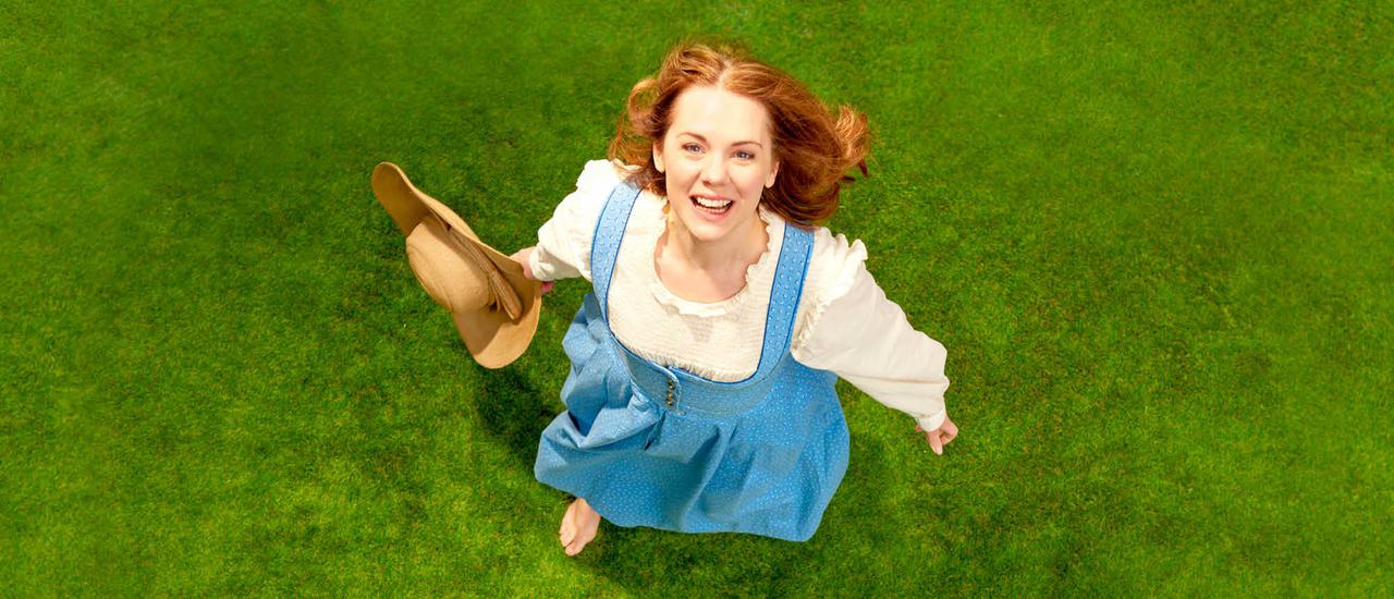 A woman in a blue dress and white dress is seen from above. She looks up at the camera smiling. Her arms are spread slightly, and she holds a straw hat in her right hand. She has red hair and bare feet and is standing on green grass.