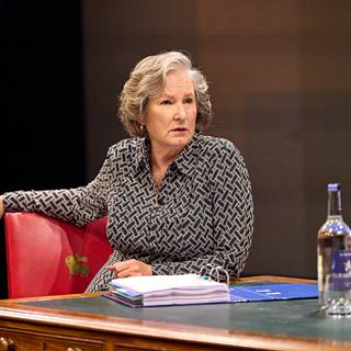 An older woman with smart grey hair sits at a desk, with one hand resting on her chair. She wears a patterned black and white dress, with a troubled expression on her face. On the desk is a large fild of papers, a bottle of water and two small glasses.