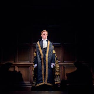 A man with red/brown hair stands on stage, wearning a long ceromonial black gown, with gold trimming. He has a slightly smug expression. On the wall above his head, the coat of arms of the United Kingdom can be seen.