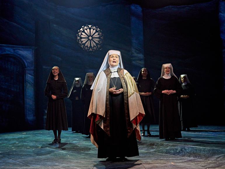 Mother Abbess (Janis Kelly) is dressed in ceremonial white robes on top of her habit, her hands together in front of her, she is looking up to the sky and signing. The other Sister's are gathered behind her also singing and we can see a round decorative window in the grey stone walls with light shining through.