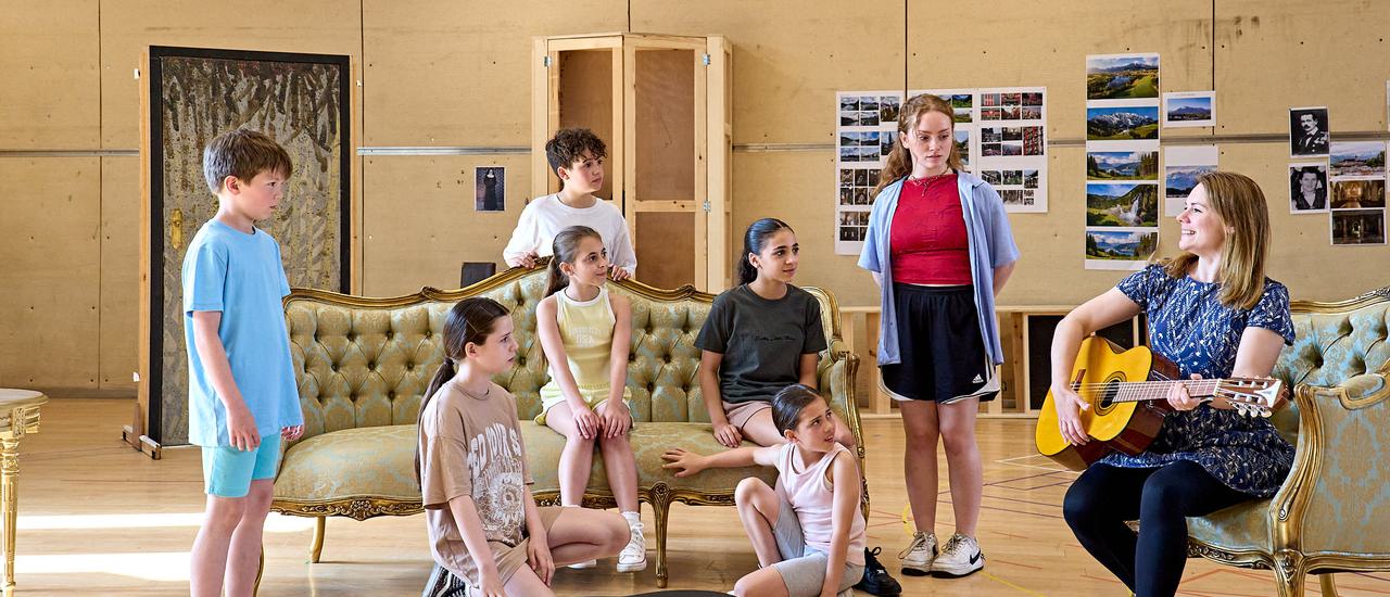 The von Trapp children are all gathered around Gina Beck who is sat in a chair holding a guitar, some of the children are sat on a pale green and wooden decorative sofa. On the floor is a black guitar case.