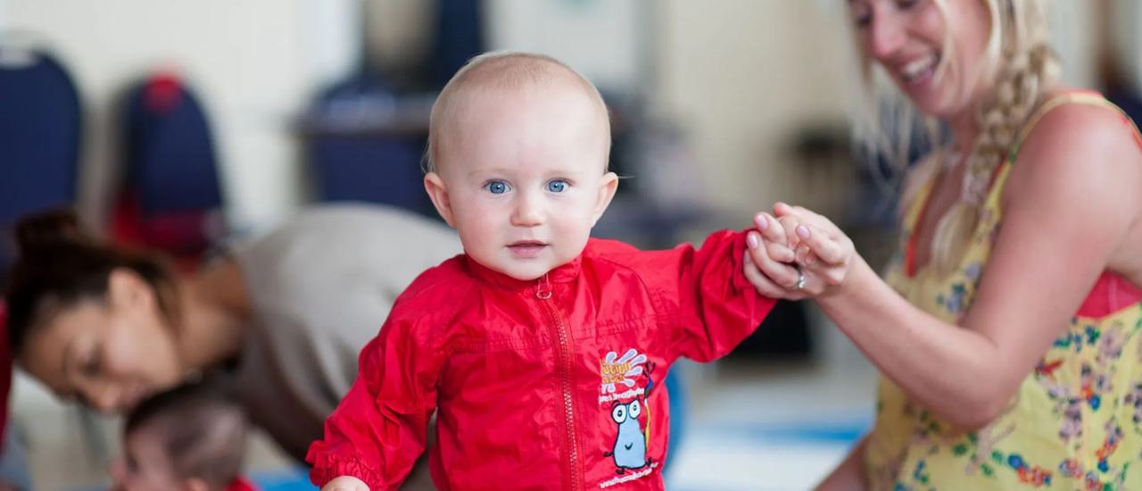 A baby in a red all in one paint suit smiles at the camera while holding a parent's hand.