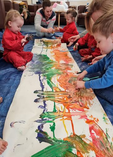 Babies paint a long strip of paper with green, orange, blue and red.