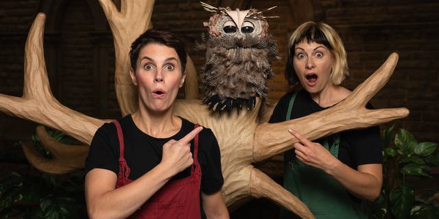 Two actors wearing dungarees stand next to a tree pointing at a Puppet owl.