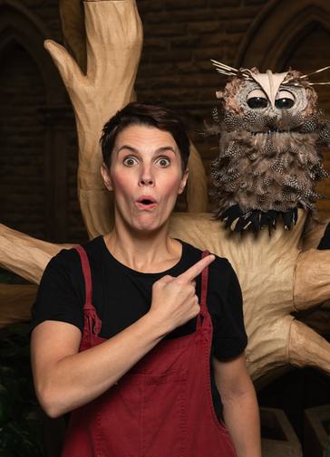 An actress pulls a silly face while pointing at an owl puppet on the left which sits in a paper tree.