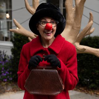 An actress is dressed as a Ladybird, with a red nose, red coat and black hat and gloves. She holds a a handbag clutched to her chest smiling pensively.