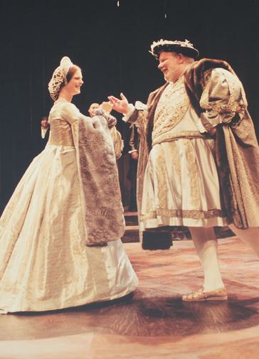 A woman and man dressed in full Tudor costume stand facing each other and smiling, their hands joined as though circling each other in a dance. The rich brocade fabrics are in shades of gold, trimmed with fur. The woman wears a high curved head-dress and a snood over her hair, while the man wears a dark hat trimmed with white feathers