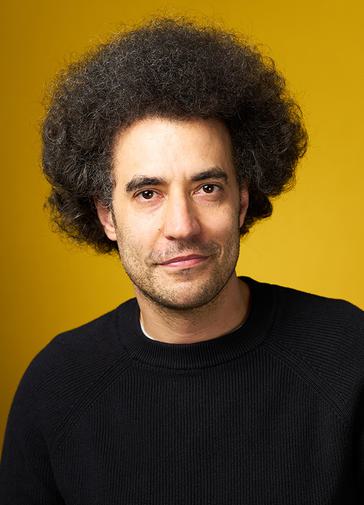 Artistic Director Justin Audibert smiles at the camera. He is wearing a black jumper and is in front of a yellow background.