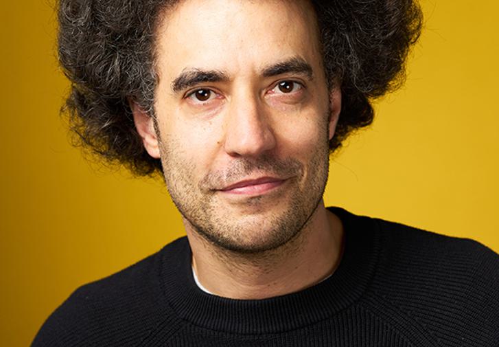 Artistic Director Justin Audibert smiles at the camera. He is wearing a black jumper and is in front of a yellow background.